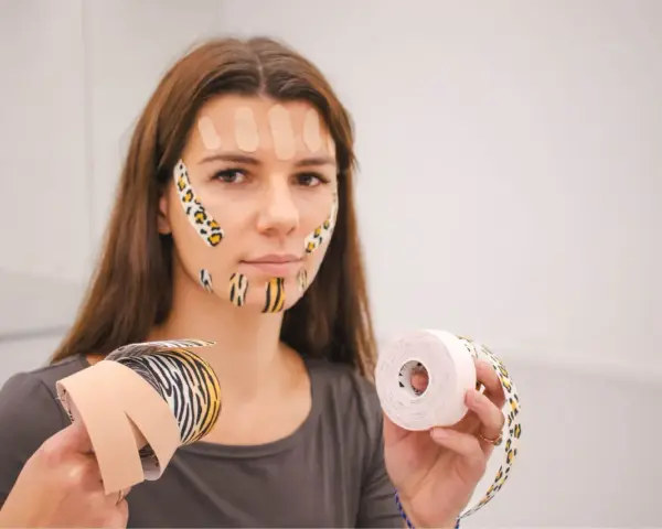 Kobieta z taśmami na twarzy trzyma rolki taśmy kinezjologicznej.