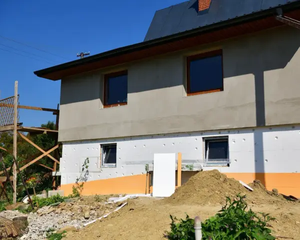Budynek w trakcie ocieplania styropianem. Widoczne okna, rusztowanie i ziemia. Trzeba uważać na **mostek termiczny** przy oknach.