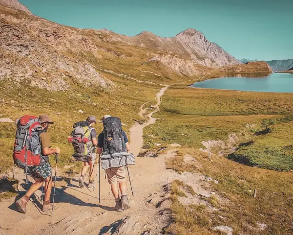 Trois randonneurs équipés pour le tour du Queyras traversent un sentier de montagne, un lac bleu et des sommets rocheux en toile de fond.