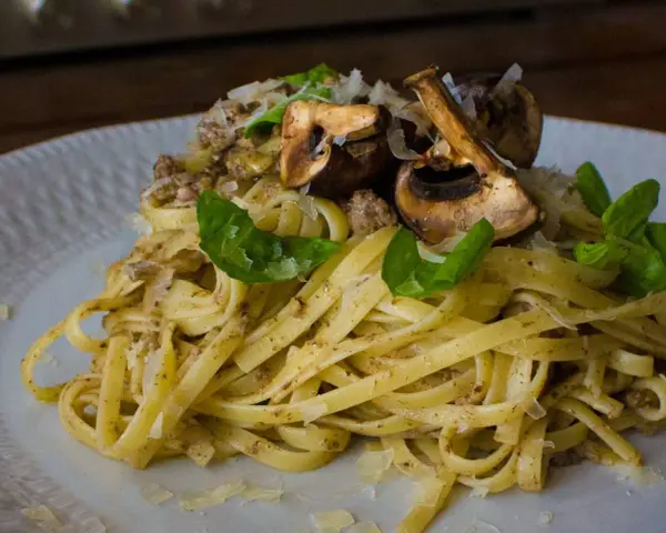 Pasta con champiñones, carne picada y queso rallado, decorada con hojas verdes.