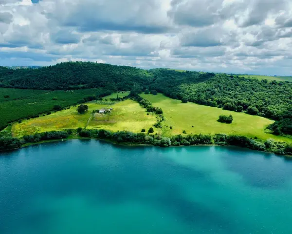 Acque turchesi del lago di Martignano lambiscono dolci colline verdi, con un casale isolato tra i campi.