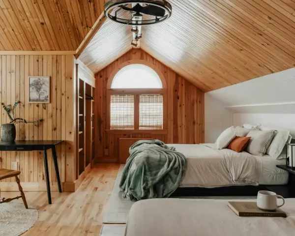 Dormitorio con paredes y techo de madera, cama con mantas y almohadas, ventana arqueada y escritorio.