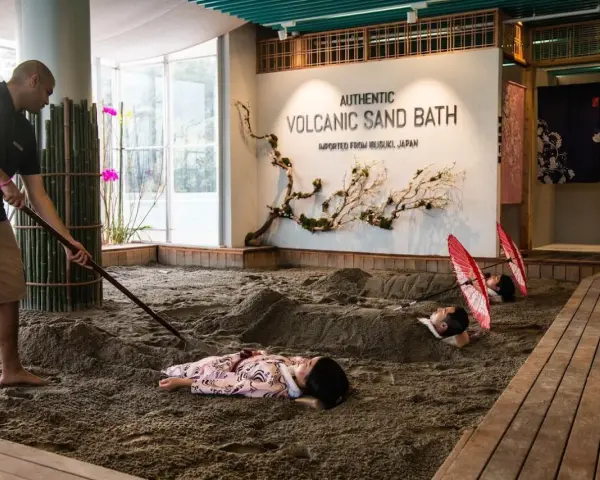 A man rakes sand for a volcanic sand bath. Two people lie buried in the sand, with red umbrellas over their heads.