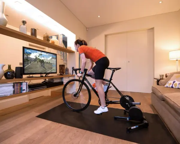 Woman in her home cycling pain cave, training indoors on a bike trainer while watching a virtual race on a large screen.