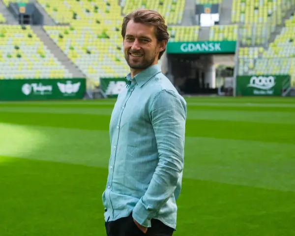 Uśmiechnięty Tomasz Kaczmarek, trener, stoi na tle stadionu w Gdańsku.