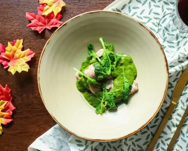 Piatto di carne con salsa verde cremosa, guarnito con cime di rapa fresche. Accanto, foglie autunnali e posate dorate.