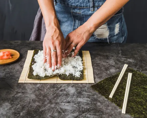 Ręce rozkładają ryż na nori, przygotowując się do zwijania sushi. To kluczowy krok, jak ugotować ryż do sushi.