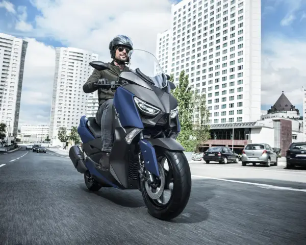 Mężczyzna w kasku i okularach przeciwsłonecznych jedzie niebieskim skuterem Yamaha X-MAX 400 po miejskiej drodze. W tle wysokie budynki i samochody.