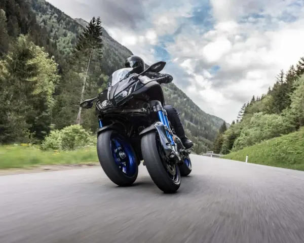 Motocyklista na Yamaha Niken pokonuje zakręt na górskiej drodze, otoczony zielenią i chmurami.