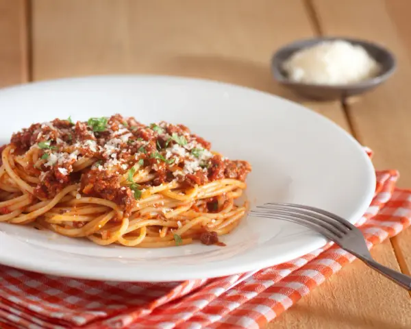 Talerz spaghetti bolognese z parmezanem i natką pietruszki, obok widelec i ser w miseczce.