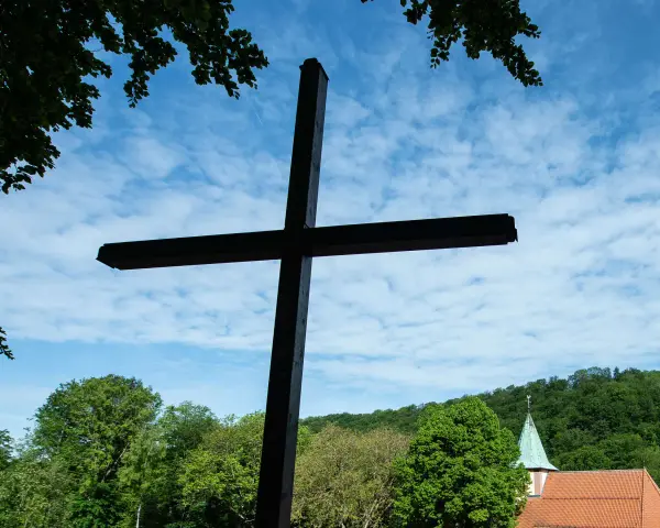 Ein großes Holzkreuz ragt vor blauem Himmel mit Wolken auf. Im Hintergrund eine Kirche und grüne Bäume. Wann ist Christi Himmelfahrt?