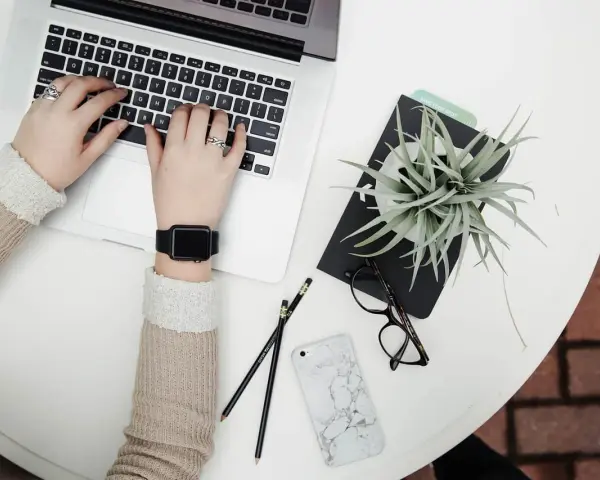 Kobieta pracuje na laptopie, obok leżą okulary, telefon i roślina. To idealne zdjęcia w chmurze do inspiracji.