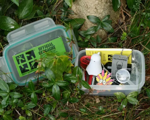 Dwa pojemniki z zawartością do geocaching co to. W jednym zielona naklejka "Official Geocache", w drugim drobne przedmioty jak duch, lupa i "Travel Bug".