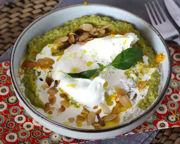 Un bol de purée verte, peut-être une sauce petit pois, garni d'un œuf poché, de crème, d'amandes effilées et d'une feuille de basilic.