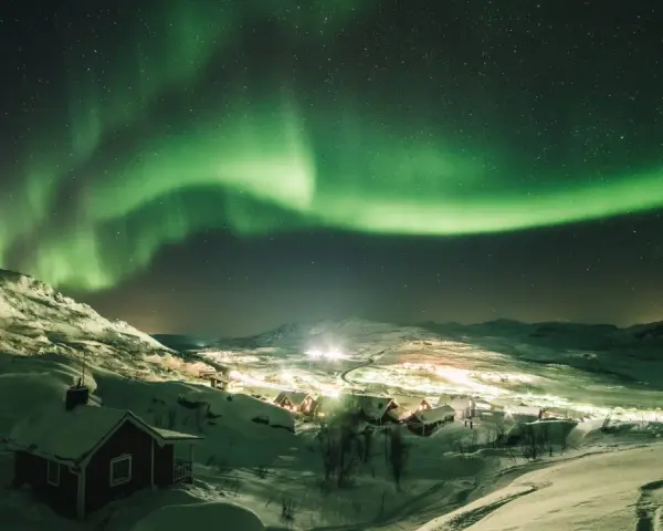 Snöklädda berg och en liten by badar i norrskenets gröna sken. En magisk natt att se norrsken.