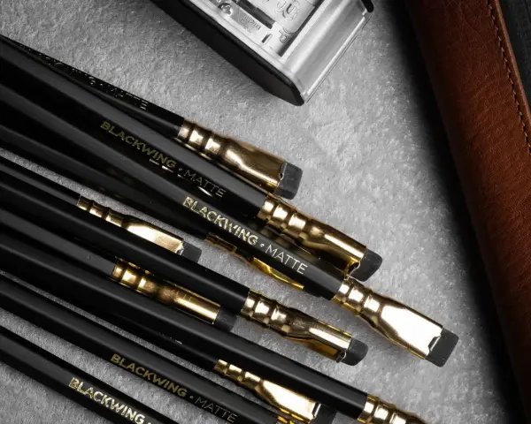 En samling svarta Blackwing-pennor med guldfattningar och suddgummin. En brun läderfodral och en metallbehållare syns också.