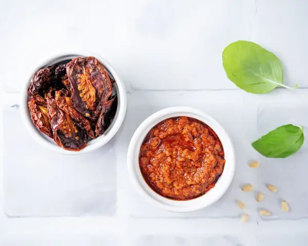 Due ciotoline bianche: una con pomodori secchi interi, l'altra con un invitante pesto di pomodori secchi. Foglie di basilico e pinoli completano la scena.