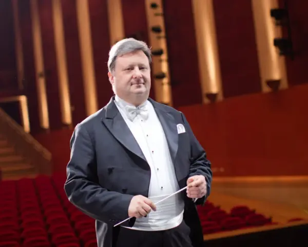 Jacek Boniecki, dyrygent w eleganckim fraku, z batutą w dłoni, na tle czerwonej widowni. Jego życiorys to historia pasji do muzyki.