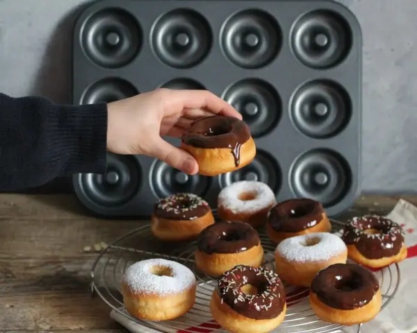 Une main tient un donut glacé au chocolat, prêt à être dégusté. Une belle collection de donuts au four, certains saupoudrés de sucre glace, d'autres de glaçage au chocolat et de vermicelles colorés, attendent sur une grille.