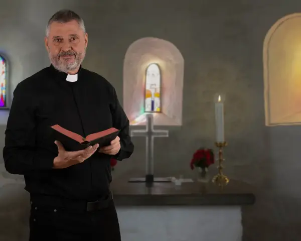 Ein Mann mit grauem Bart und Priesterkragen hält ein Buch in einer Kirche. Er hat sich entschieden, Priester zu werden.