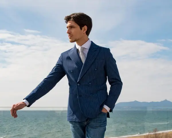 Uomo con giacca doppiopetto blu a righe, camicia bianca e cravatta, jeans. Eleganza classica uomo con vista mare.