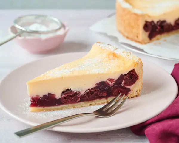 Ein saftiges Stück Kirsch Schmand Kuchen mit cremigem Belag und Kirschen auf Mürbeteigboden, bestäubt mit Puderzucker.