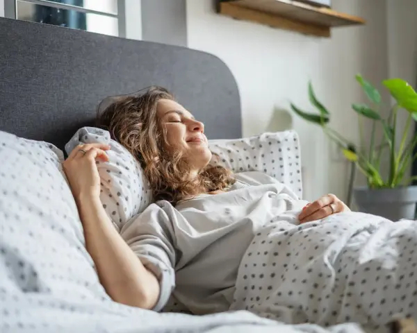 Une femme souriante se repose dans un lit douillet. Elle semble heureuse, une belle illustration de comment choisir une bonne literie pour un sommeil réparateur.