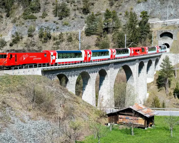 The red and white Glacier Express train travels across a stone viaduct, a scenic journey from Chur to Zermatt.