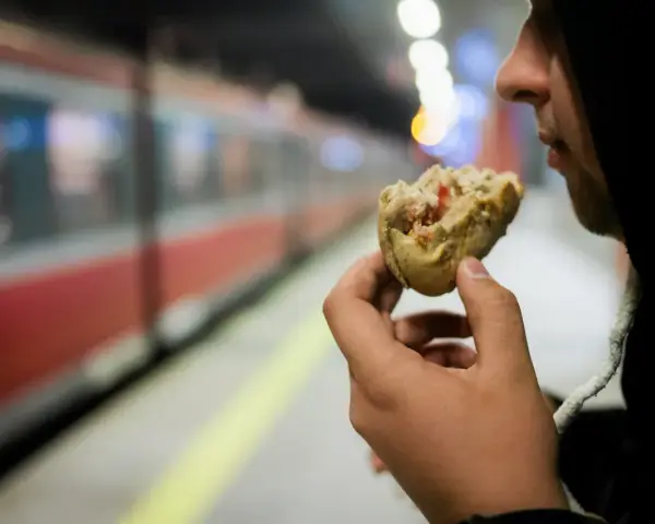 Osoba w kapturze je kanapkę na tle pociągu na peronie.