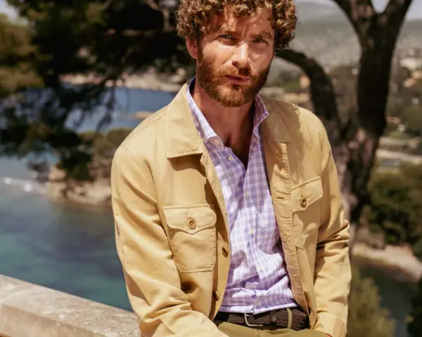 Un uomo con barba e capelli ricci indossa una camicia a quadri viola e una giacca color sabbia. Ottima idea su cosa mettere sopra la camicia uomo per un look casual chic.