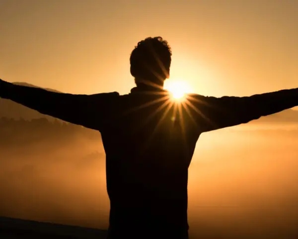 Silhouette eines Mannes mit ausgebreiteten Armen im Sonnenaufgang. Die Sonne bricht durch seine Silhouette und wirft Strahlen. Ein Symbol für Hoffnung und wie wird man Christ.