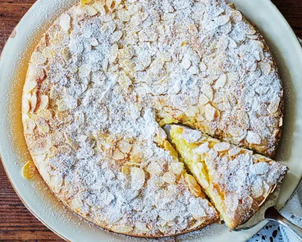 En saftig mandelkaka med citron, toppad med flagad mandel och pudrad florsocker. En bit är uppskuren.