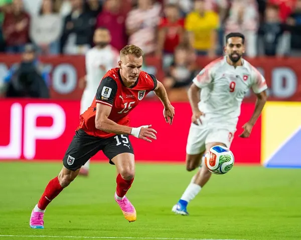 Jugadores de fútbol en acción. El número 15 de Austria corre con el balón, mientras otro jugador se acerca. ¿Quién juega hoy?