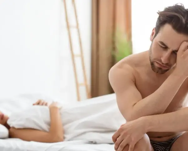 Un homme pensif, le regard baissé, semble préoccupé par un besoin sexuel non satisfait, tandis qu'une femme dort paisiblement à ses côtés.