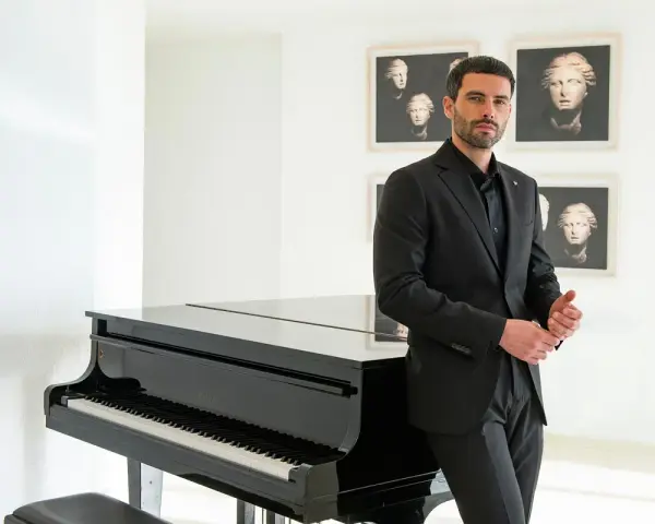 Uomo con elegante outfit per andare a teatro, in completo nero, accanto a un pianoforte a coda.