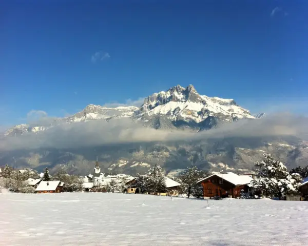 Un pueblo alpino cubierto de nieve, con casas de madera y una iglesia, bajo imponentes montañas nevadas. Un paisaje de invierno en francés.