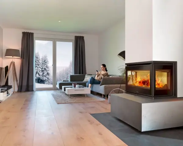 Mujer en sofá mira móvil junto a chimenea de cassette pellets con fuego vivo. Ventana muestra paisaje nevado.