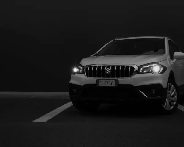 Białe Suzuki SX4 na ciemnym tle, z włączonymi światłami.