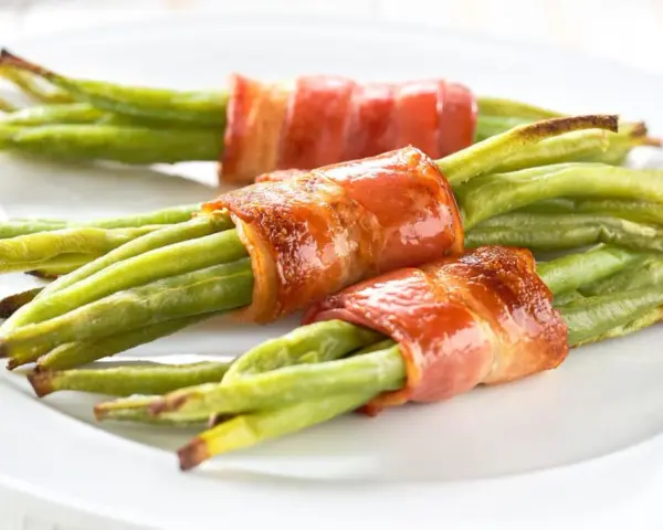 Drei Portionen Bohnen im Speckmantel auf einem weißen Teller. Der Speck ist knusprig gebraten und umhüllt die grünen Bohnen.