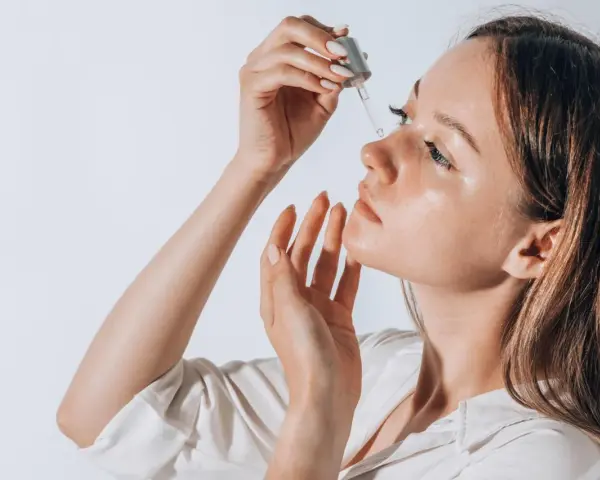 Frauenhand trägt Tropfen eines Serums auf ihr Gesicht auf. Ob Squalan oder Hyaluron, die Haut strahlt Feuchtigkeit aus.