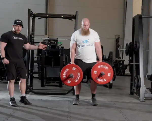 En man med skägg utför farmers walk med tunga vikter. En tränare visar hur övningen ska göras.