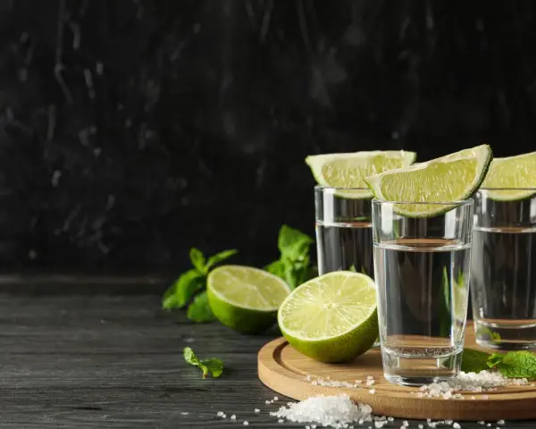 Trzy kieliszki z drinkiem z mezcalem lub tequilą, ozdobione ćwiartkami limonki i miętą, na tle czarnej ściany.