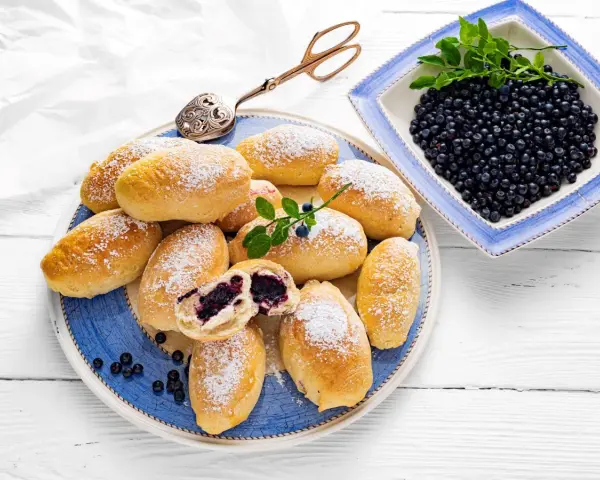 Pyszne jagodzianki posypane cukrem pudrem, obok miseczka pełna świeżych jagód. Idealne na letnie popołudnie.