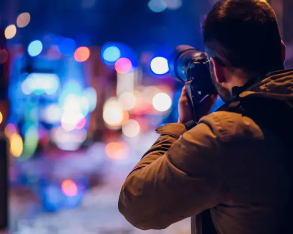 Mężczyzna z aparatem w ręku, uchwycony w trakcie nocnej fotografii. Rozmyte, kolorowe światła miasta tworzą magiczne tło.