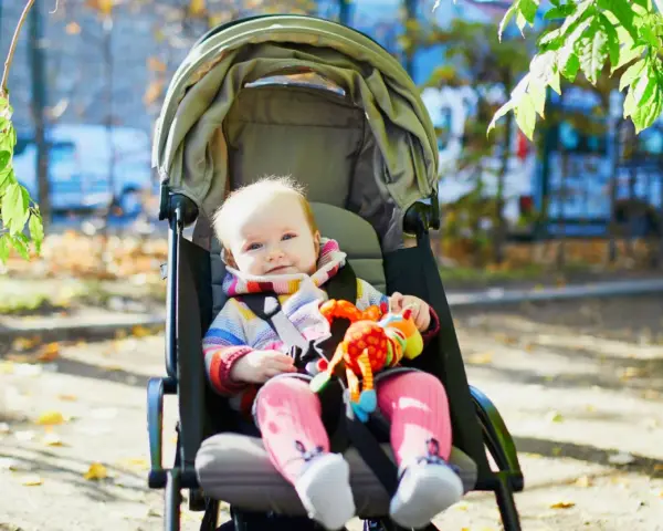 Uśmiechnięte dziecko w kolorowym sweterku i różowych skarpetkach siedzi w spacerówce, trzymając zabawkę. Czas na spacer!