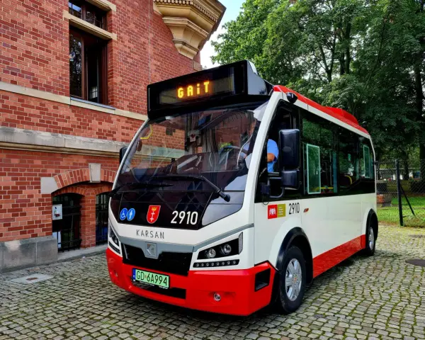 Nowy autobus 100 z napisem "GAIT" na wyświetlaczu, zaparkowany na brukowanej ulicy obok ceglanego budynku.