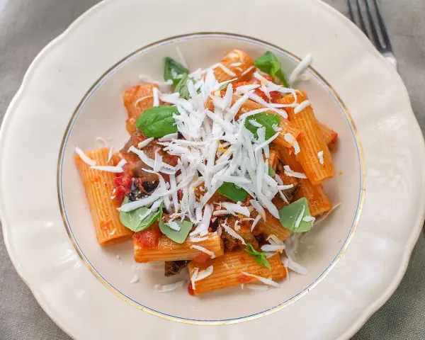 En tallrik med pasta alla norma, toppad med riven ost och färsk basilika.