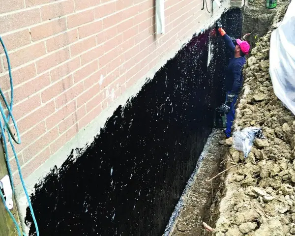 Prace przy izolacji fundamentów w starym domu. Pracownik nakłada czarną masę izolacyjną na ścianę fundamentową.