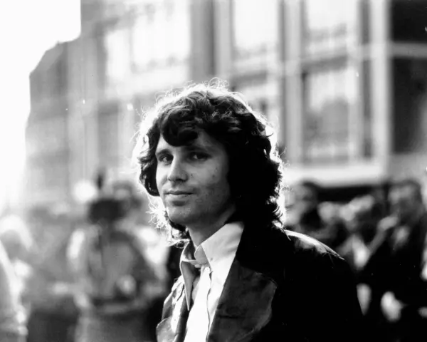 Jim Morrison, mit seinem charakteristischen lockigen Haar und einem nachdenklichen Blick, steht im Gegenlicht.