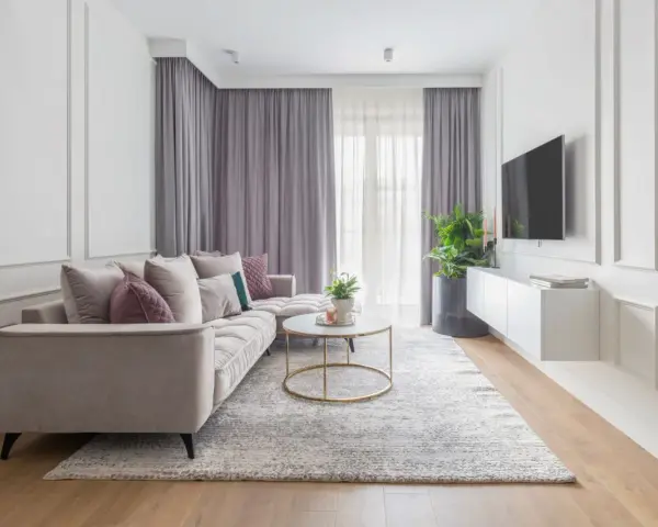 Elegancki salon w stylu modern classic. Jasna sofa, okrągły stolik kawowy, dywan i zasłony tworzą przytulną atmosferę.